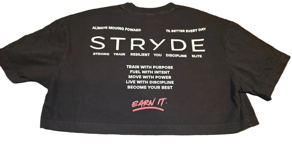 A black cropped t-shirt laid flat on a wooden floor, showing the back with white motivational text and a pink 'EARN IT.' logo.
