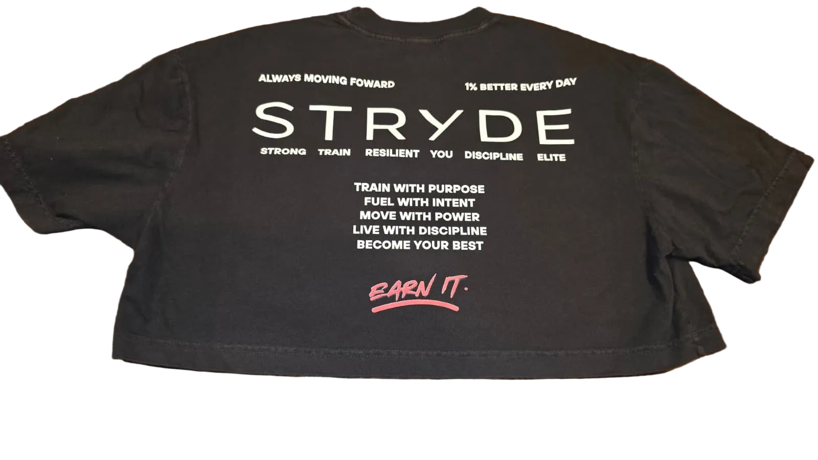 A black cropped t-shirt laid flat on a wooden floor, showing the back with white motivational text and a pink 'EARN IT.' logo.
