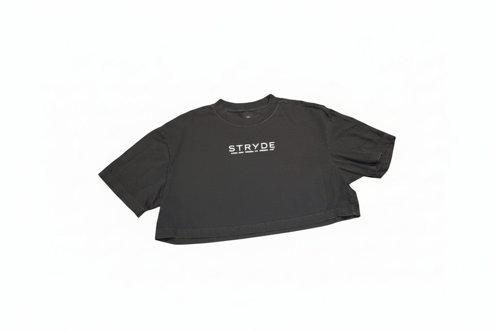 A black short-sleeve crop top t-shirt with the word 'STRYDE' printed in white across the chest, laid flat on a polished wooden table.
