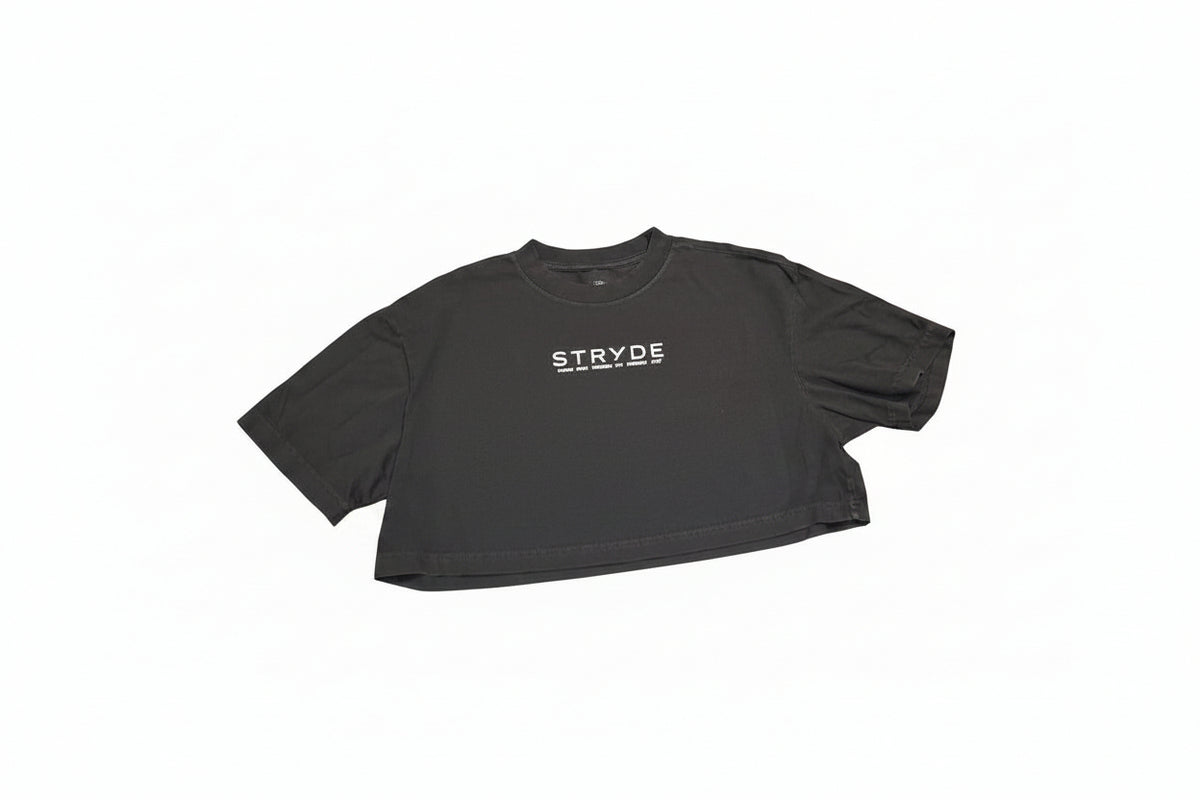 A black short-sleeve crop top t-shirt with the word 'STRYDE' printed in white across the chest, laid flat on a polished wooden table.