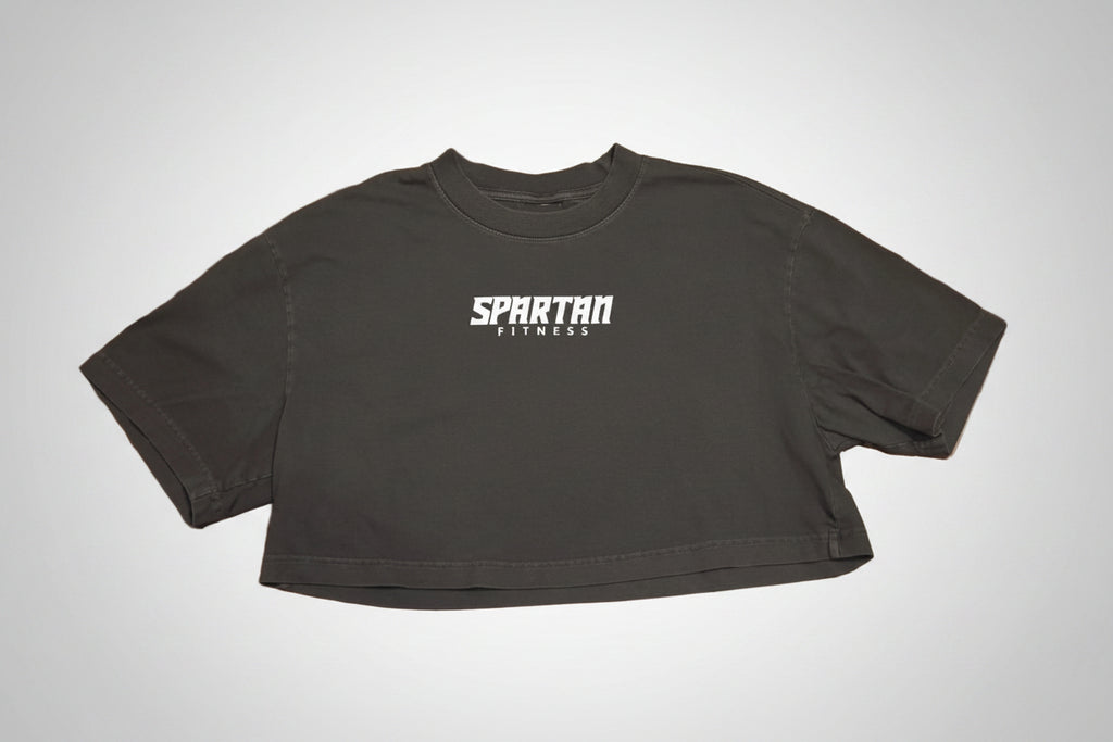 A black cropped t-shirt with 'SPARTAN FITNESS' printed in white across the chest, laid flat on a polished brown wood parquet floor.
