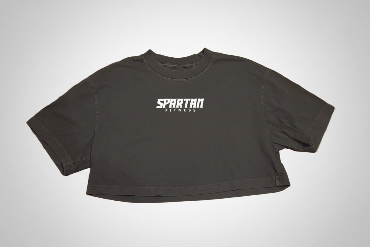 A black cropped t-shirt with 'SPARTAN FITNESS' printed in white across the chest, laid flat on a polished brown wood parquet floor.