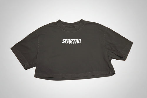 A black cropped t-shirt with 'SPARTAN FITNESS' printed in white across the chest, laid flat on a polished brown wood parquet floor.